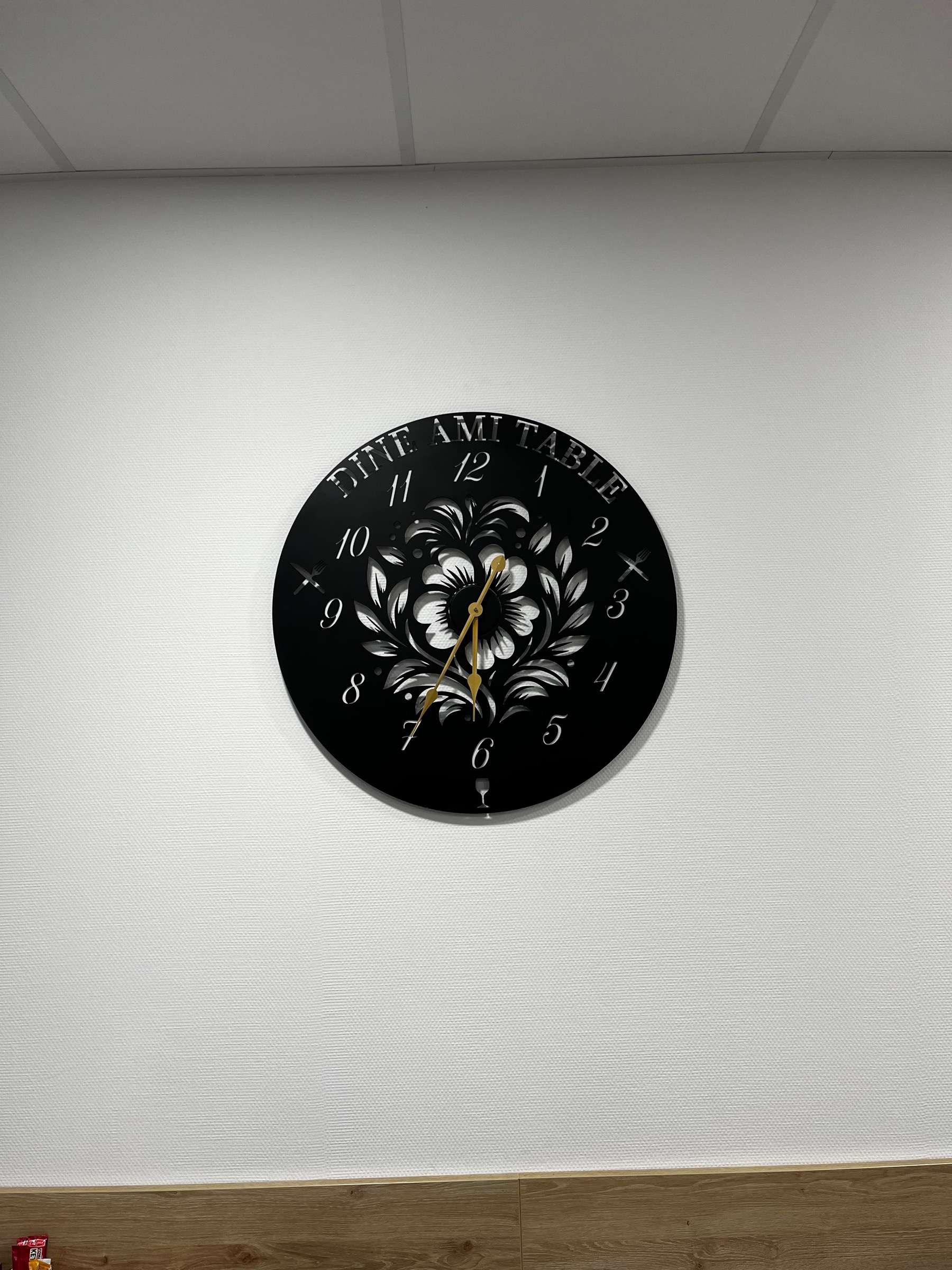 Horloge ronde en acier noir, chiffres et motif floral découpés, fixée sur un mur blanc.