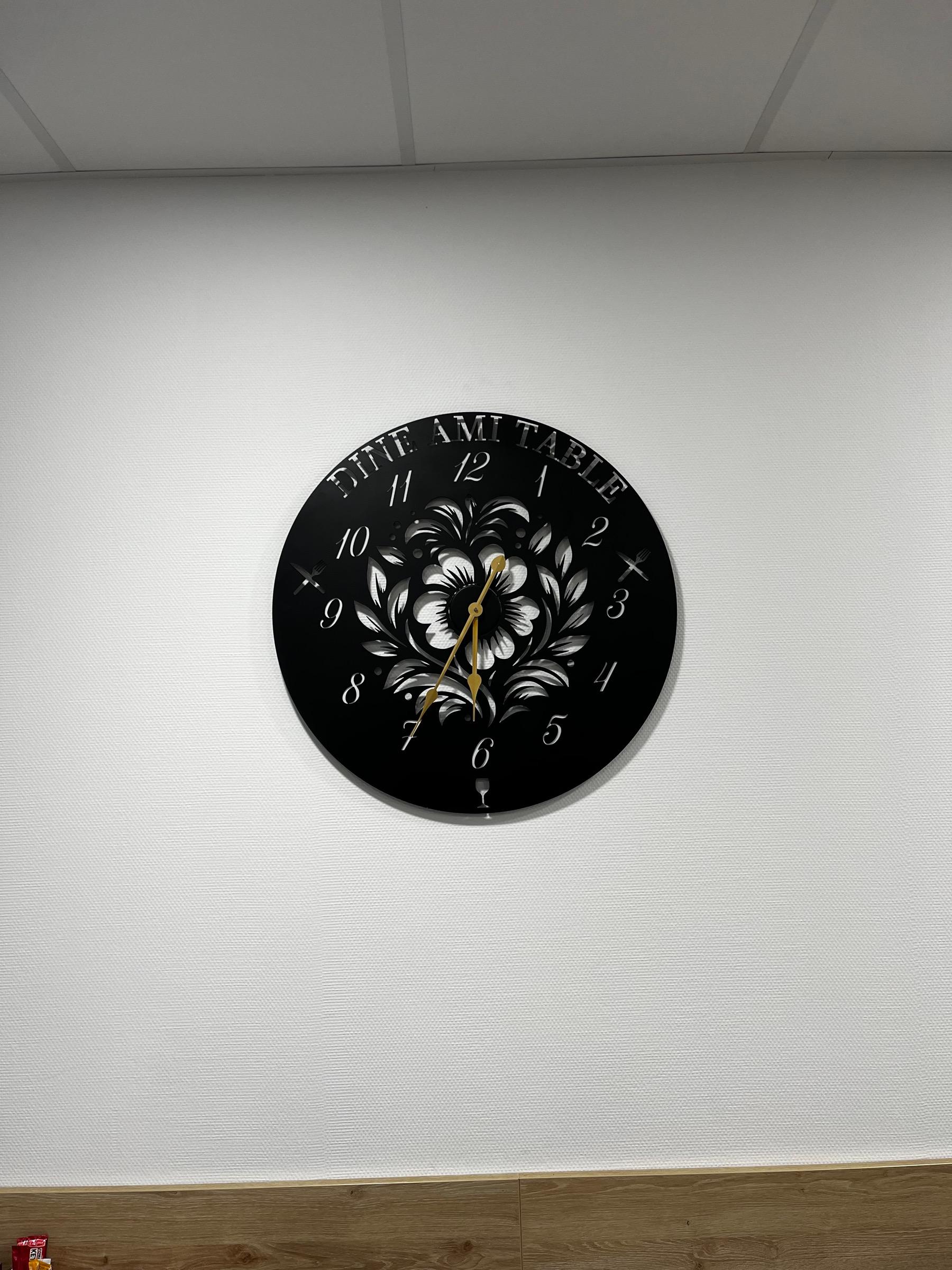 Horloge ronde en acier noir, chiffres et motif floral découpés, fixée sur un mur blanc.